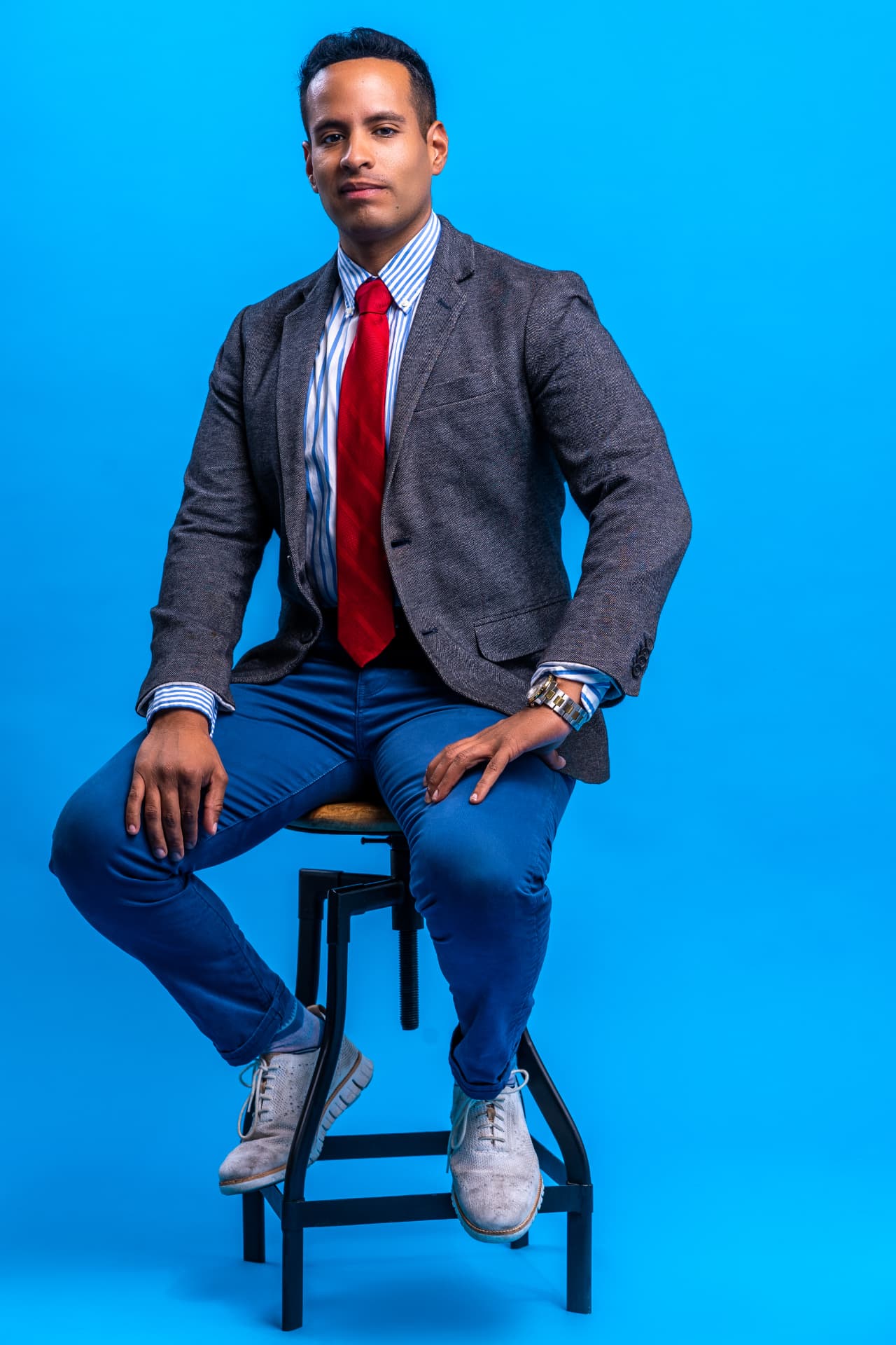 Andrew Goldstein seated on a stool against a blue backdrop in a grey jacket and red tie